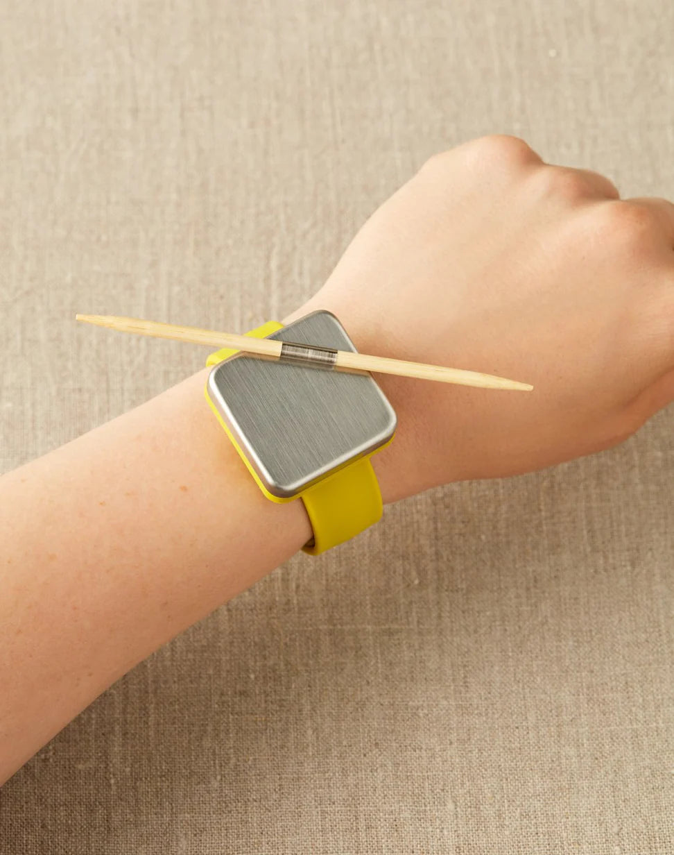 A wrist wearing a notions holder on a bracelet with a yellow strap on a beige background