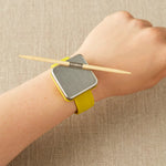 A wrist wearing a notions holder on a bracelet with a yellow strap on a beige background