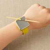 A wrist wearing a notions holder on a bracelet with a yellow strap on a beige background