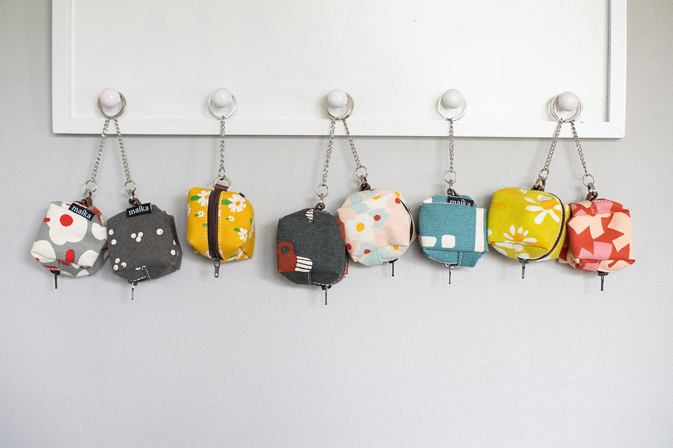 Six small fabric zip pouches in assorted colors and patterns hang from silver chains on white wall hooks against a light gray wall.