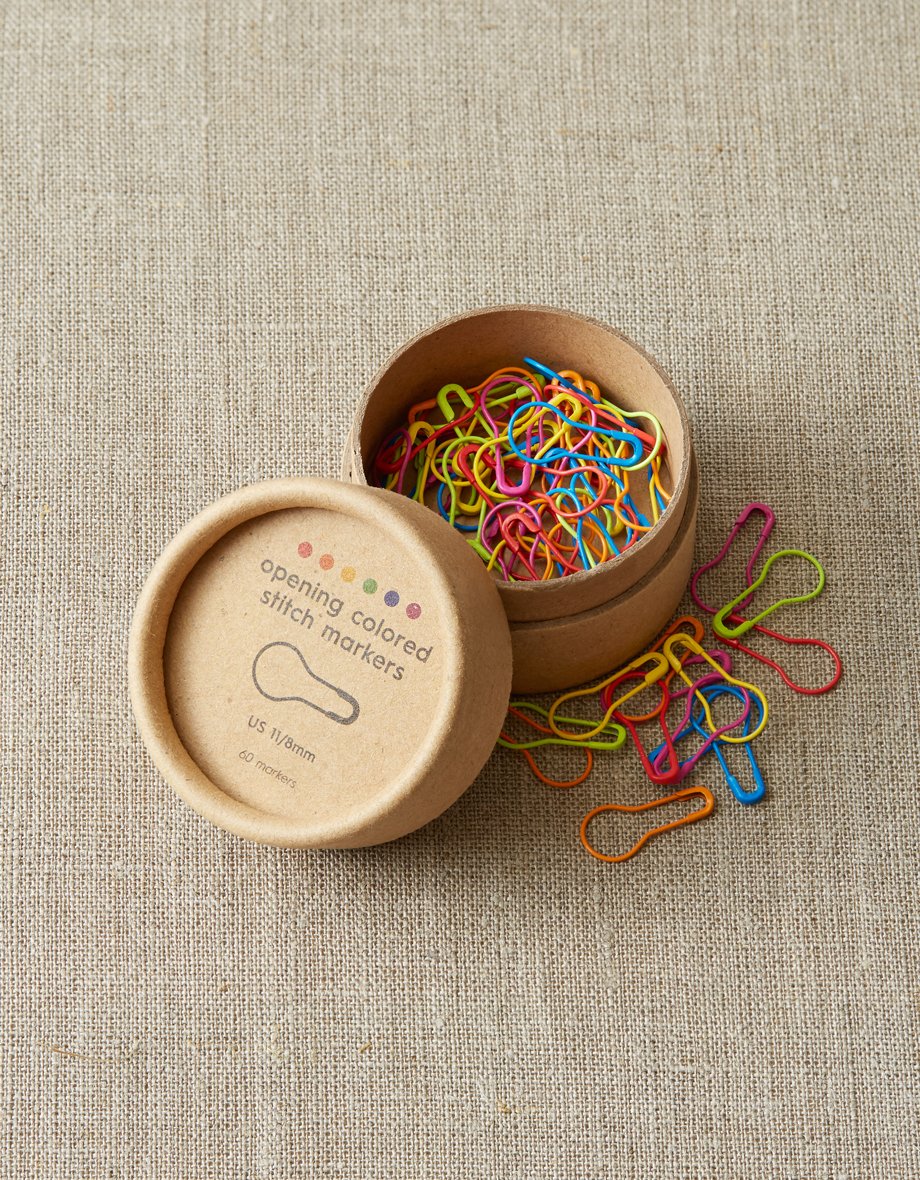 Colorful opening stitch markers in a cardboard box on a textured beige surface