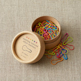 Colorful opening stitch markers in a cardboard box on a textured beige surface