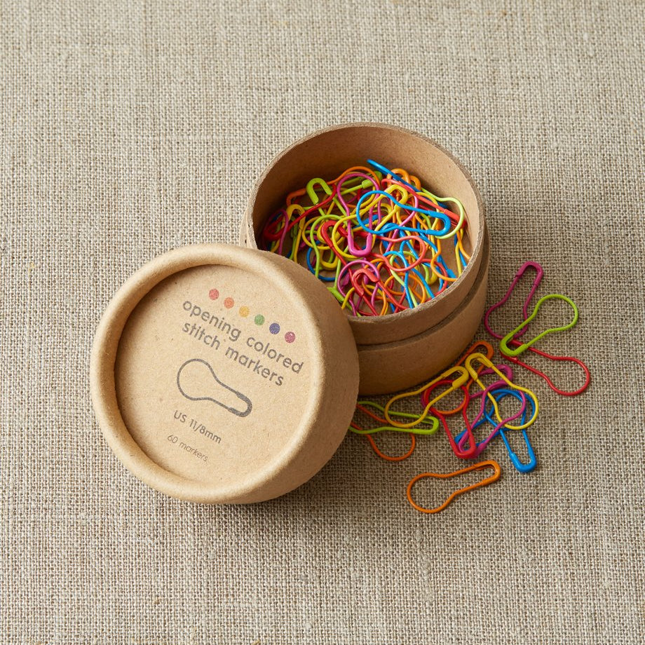 Colorful opening stitch markers in a cardboard box on a textured beige surface