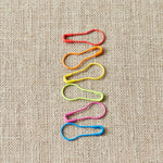 Colorful safety pin stitch markers arranged in a pattern on a textured beige background