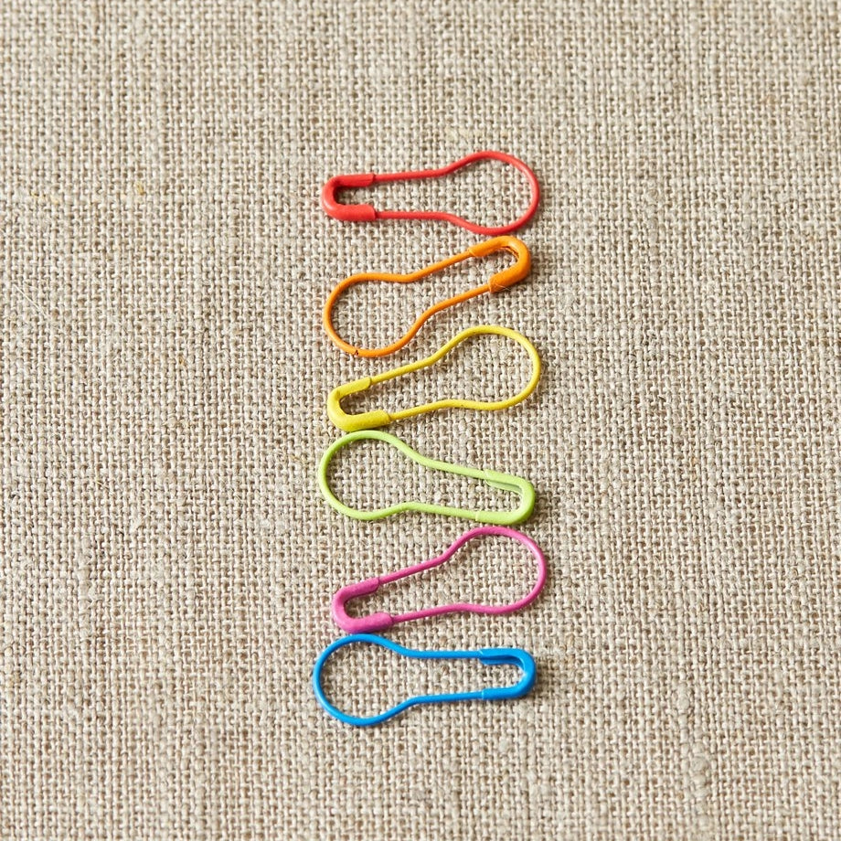 Colorful safety pin stitch markers arranged in a pattern on a textured beige background