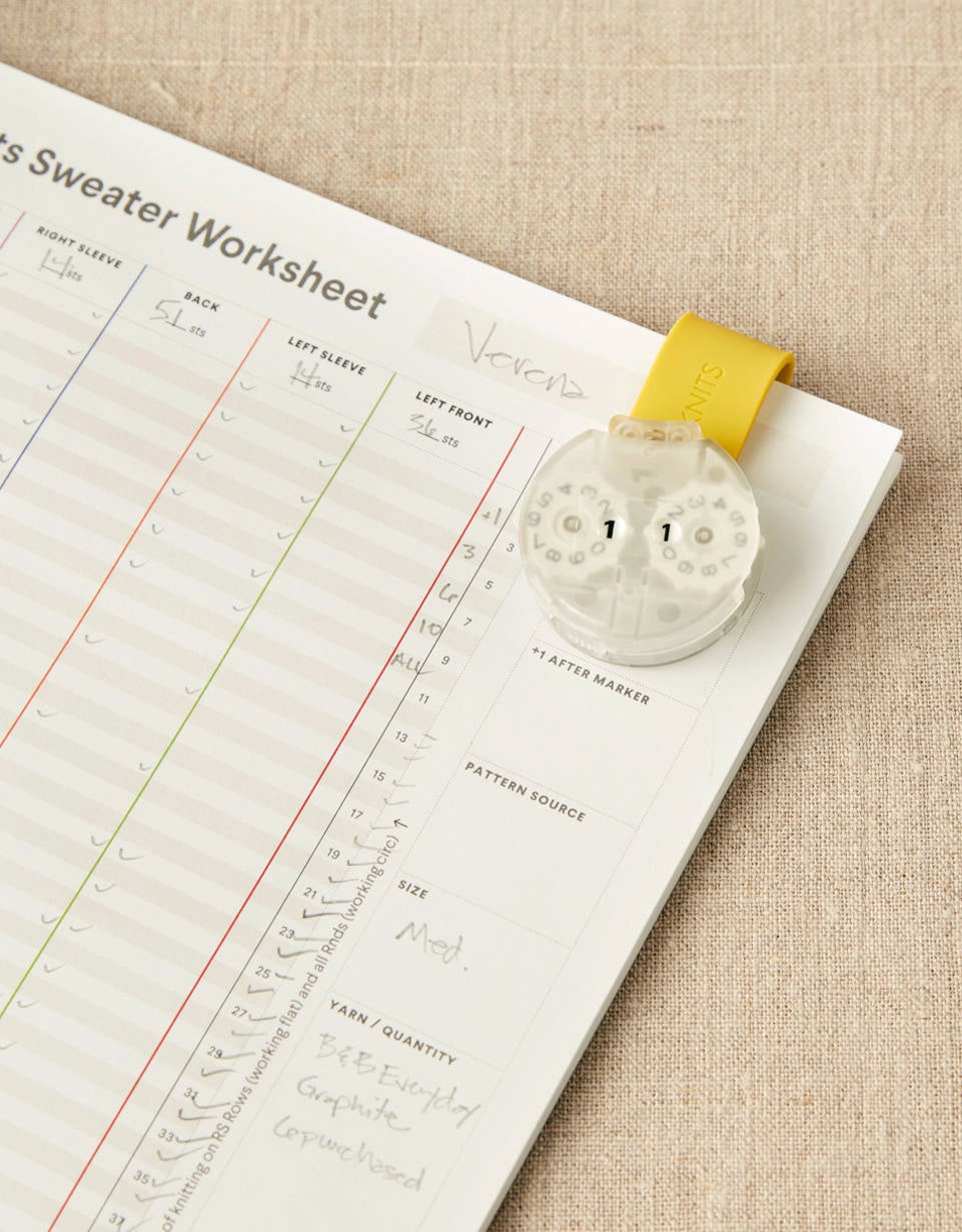 Pattern worksheet with a row counter held together with a magnetic clip. on a beige fabric background
