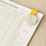 Pattern worksheet with a row counter held together with a magnetic clip. on a beige fabric background