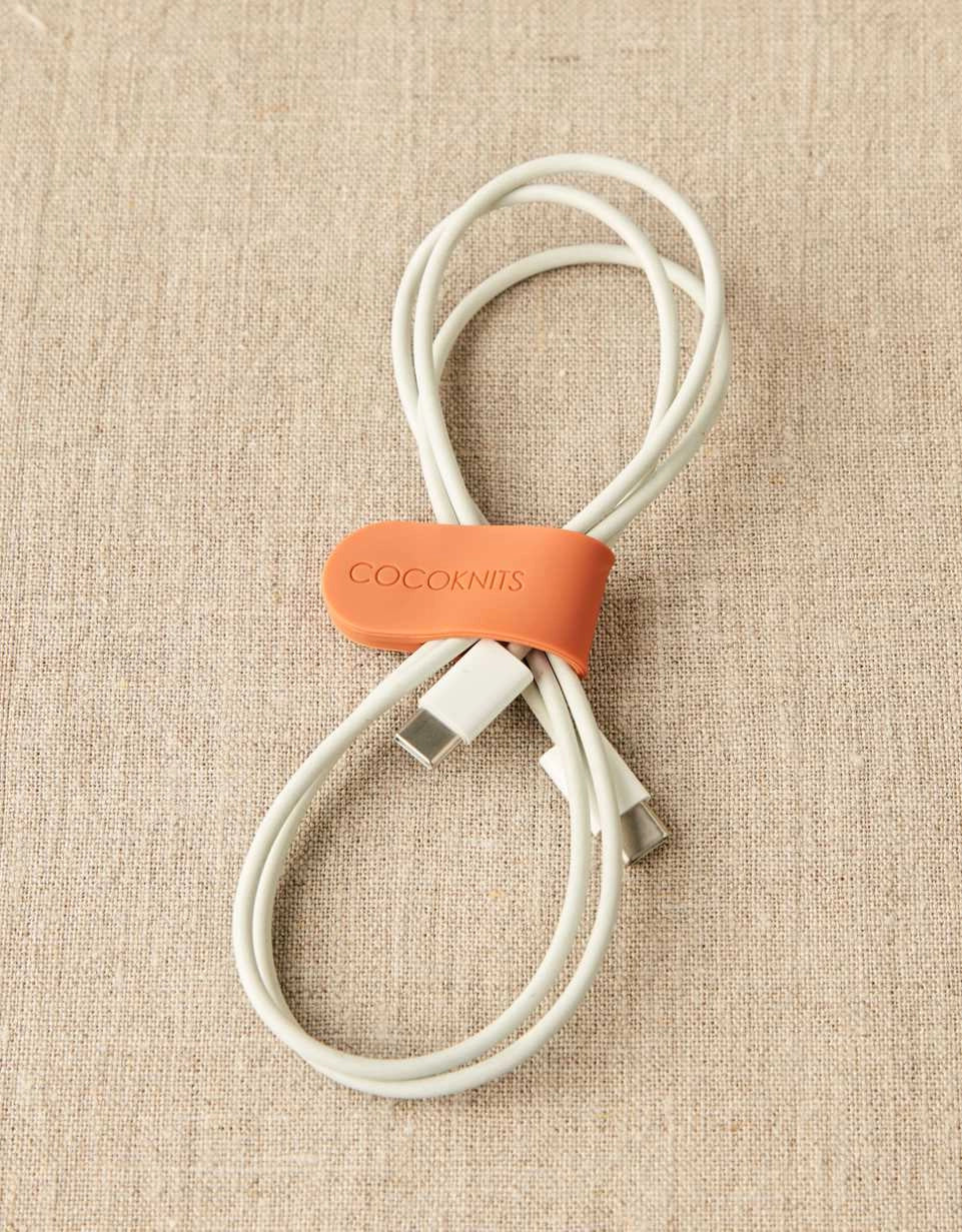 White USB cable with an orange magnetic clip labeled 'Cocoknits' on a beige fabric background
