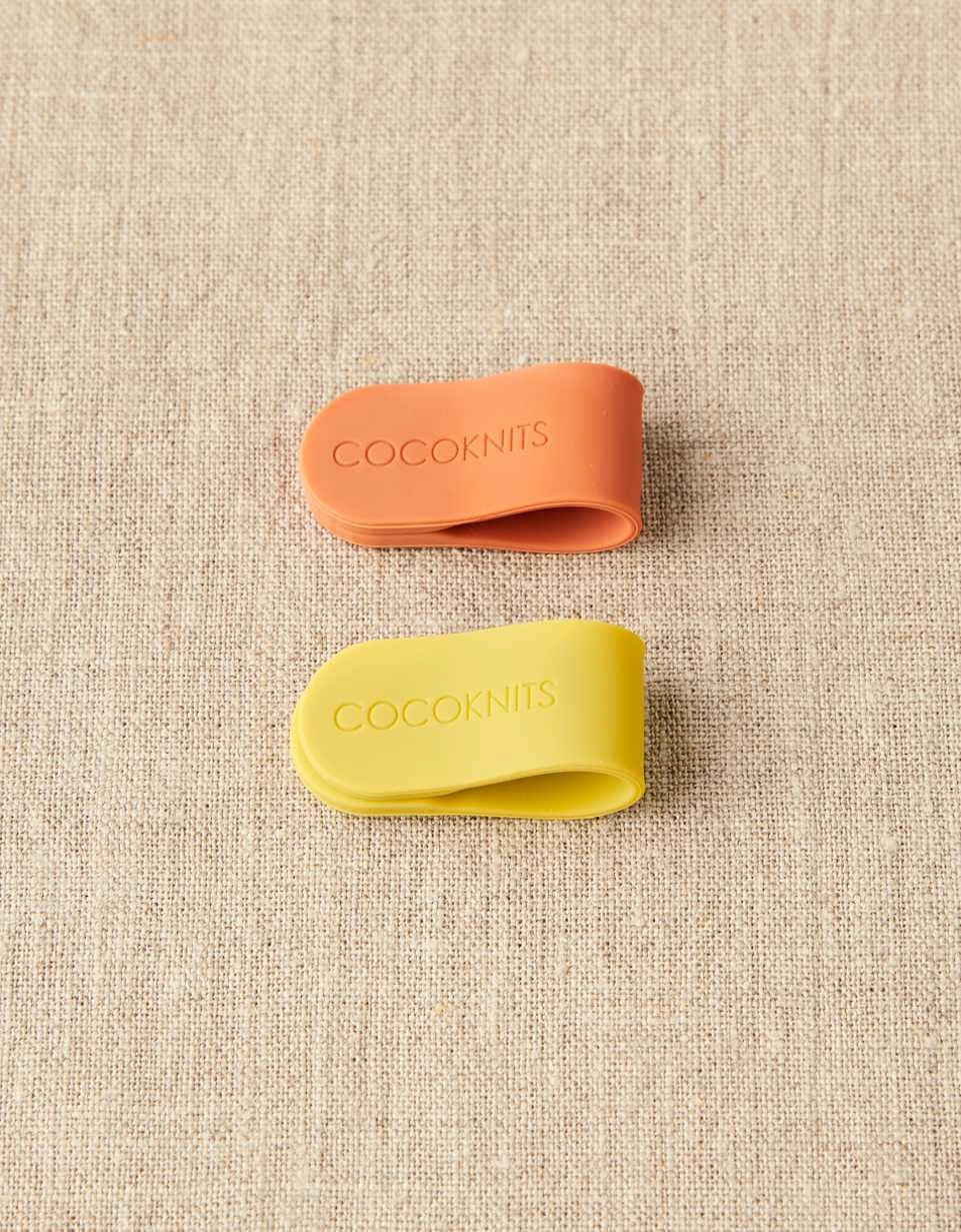 Two silicone clips, one orange and one yellow, with 'Cocoknits' branding on a textured beige background.