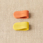 Two silicone clips, one orange and one yellow, with 'Cocoknits' branding on a textured beige background.