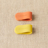 Two silicone clips, one orange and one yellow, with 'Cocoknits' branding on a textured beige background.