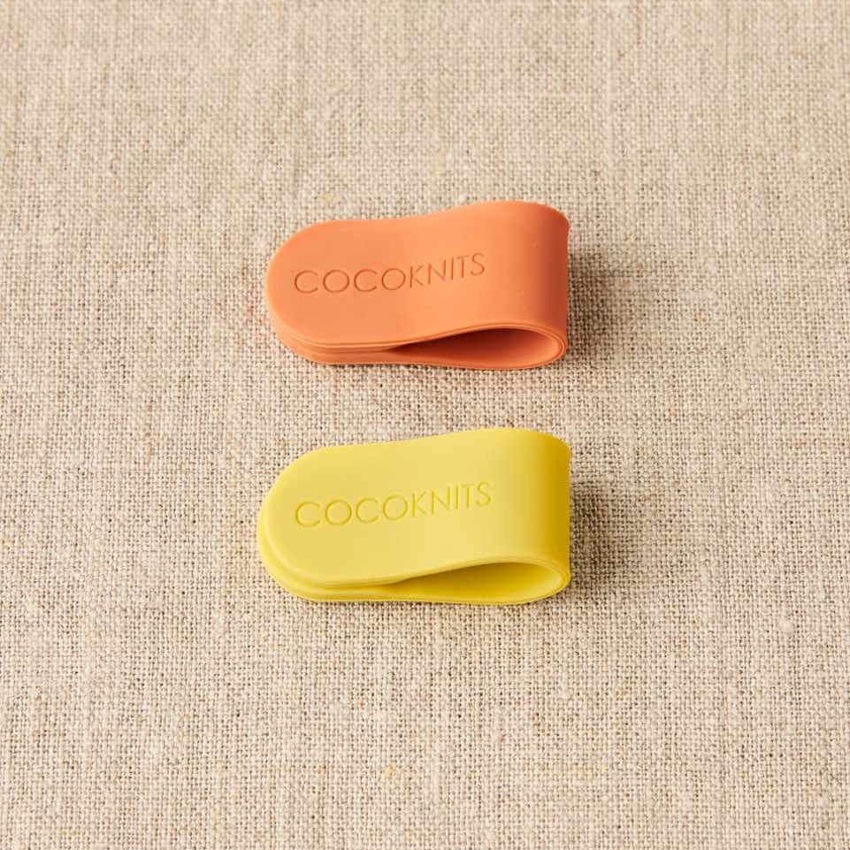 Two silicone clips, one orange and one yellow, with 'Cocoknits' branding on a textured beige background.
