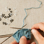Person knitting with blue yarn and black needles, using extra-small triangle stitch markers on a beige fabric background.