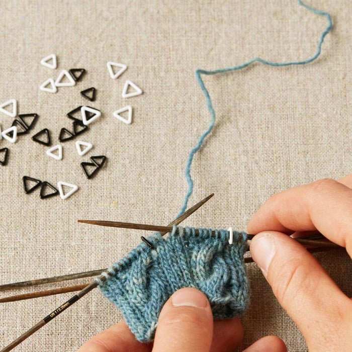 Person knitting with blue yarn and black needles, using extra-small triangle stitch markers on a beige fabric background.