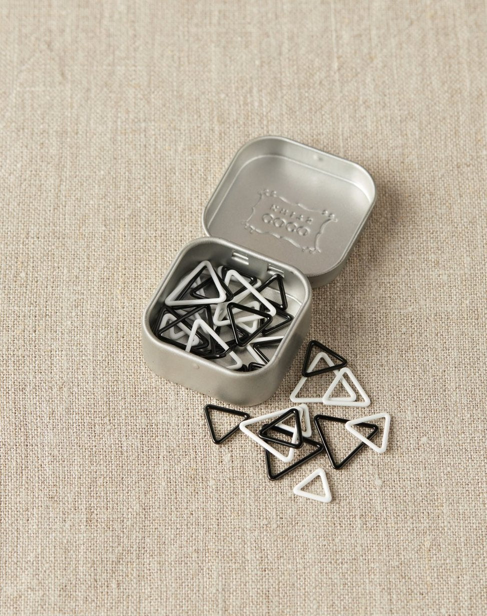 Set of triangle-shaped black and white stitch markers in a metal tin on a beige fabric background