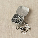 Set of triangle-shaped black and white stitch markers in a metal tin on a beige fabric background
