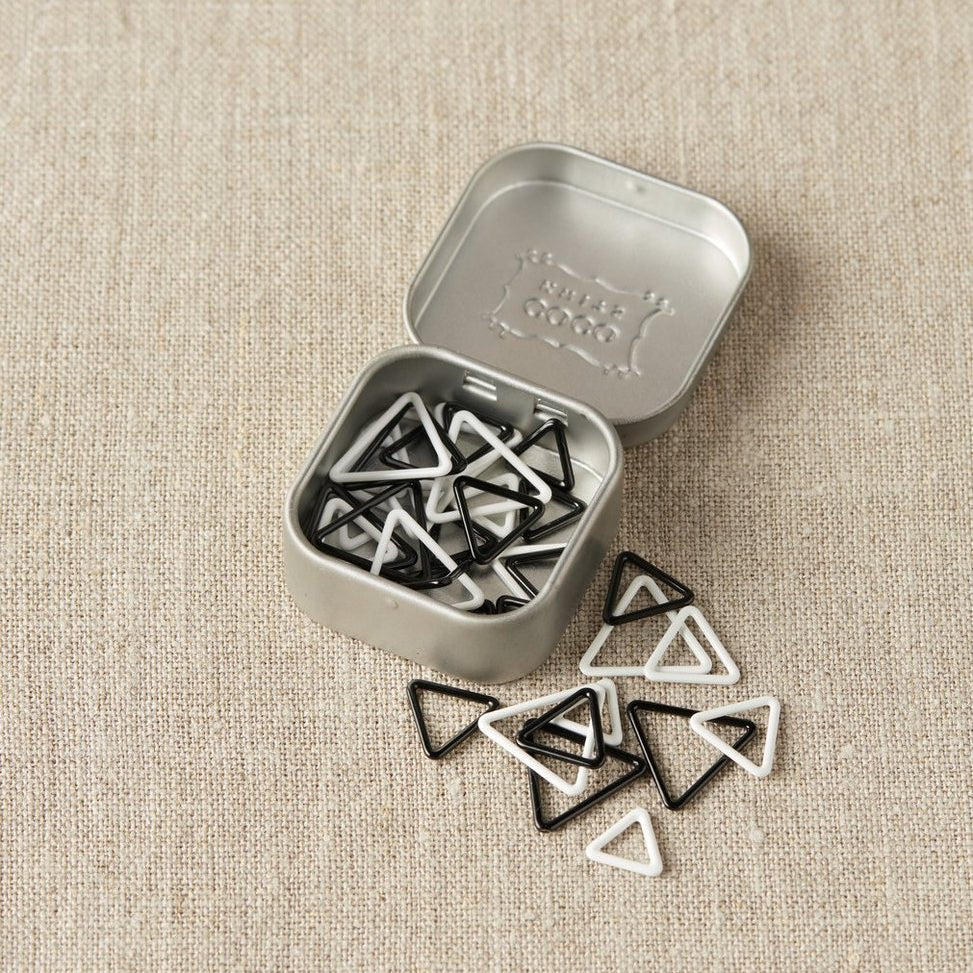 Set of triangle-shaped black and white stitch markers in a metal tin on a beige fabric background