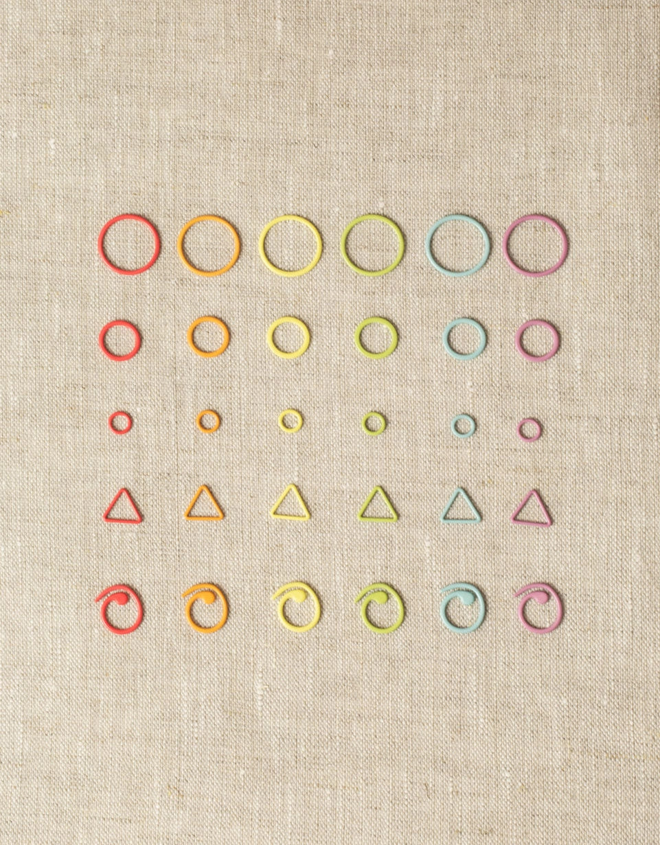 Various types of colorful stitch markers on a beige fabric background. Includes small, medium & large closed ring, triangle, & split ring stitch markers.