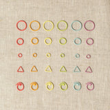 Various types of colorful stitch markers on a beige fabric background. Includes small, medium & large closed ring, triangle, & split ring stitch markers.