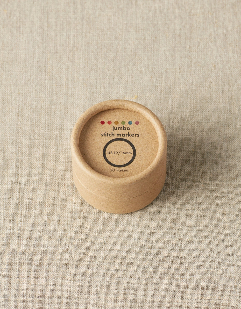 Round wooden container with 'jumbo stitch markers' label on a beige fabric background