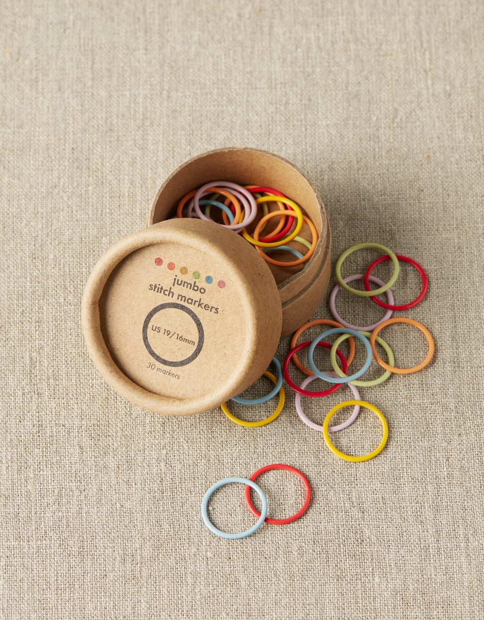Colorful jumbo stitch markers spilling out of a cardboard container on a beige fabric background