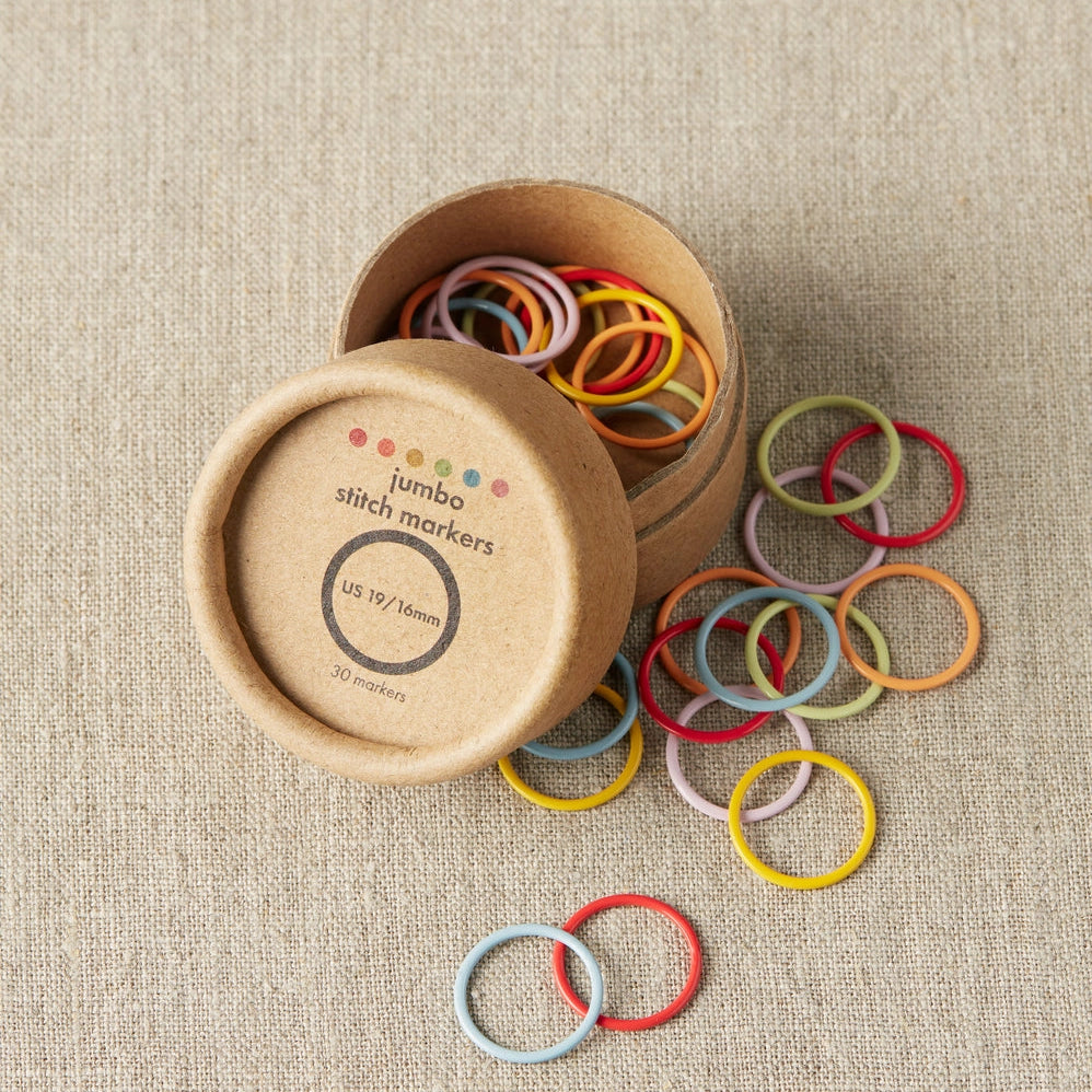 Colorful jumbo stitch markers spilling out of a cardboard container on a beige fabric background