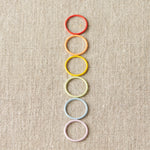 Colorful ring stitch markers arranged vertically on a textured beige background