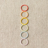 Colorful ring stitch markers arranged vertically on a textured beige background