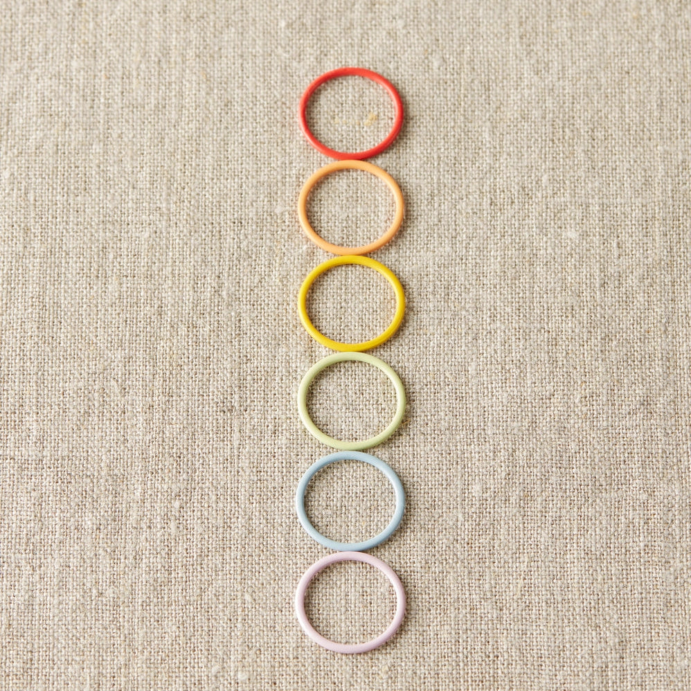 Colorful ring stitch markers arranged vertically on a textured beige background