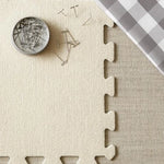 Beige interlocking foam puzzle mat with a container of pins on a checkered fabric background