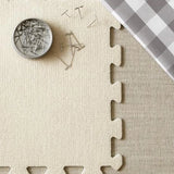 Beige interlocking foam puzzle mat with a container of pins on a checkered fabric background