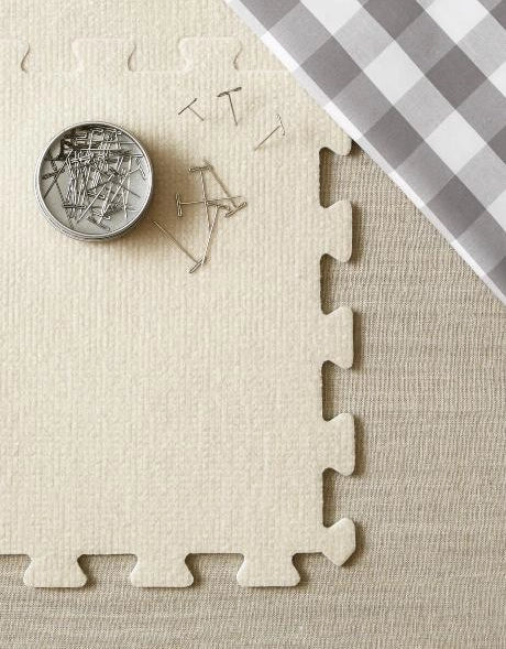 Beige interlocking foam puzzle mat with a container of pins on a checkered fabric background