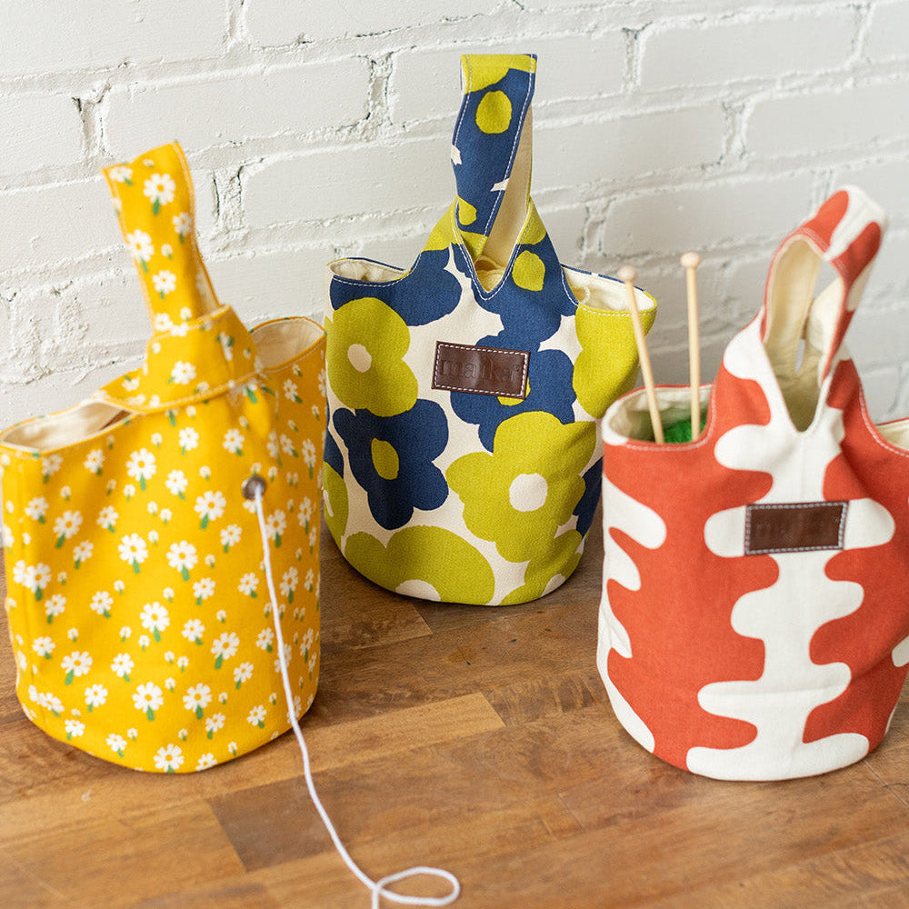 Three fabric storage bins with handles, decorated in yellow daisies, blue and yellow flowers, and red and white abstract patterns, arranged on a wooden surface against a white brick wall.