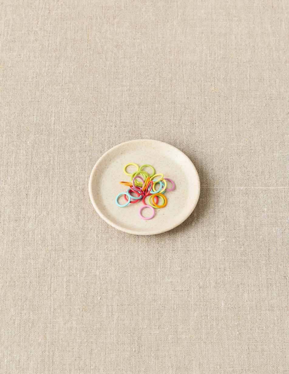 Small white plate with colorful stitch markers on a beige fabric background