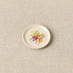 Small white plate with colorful stitch markers on a beige fabric background