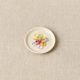 Small white plate with colorful stitch markers on a beige fabric background
