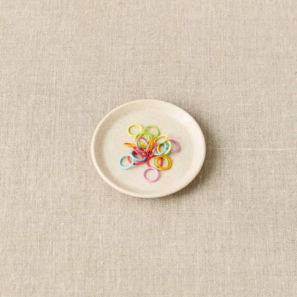 Small white plate with colorful stitch markers on a beige fabric background