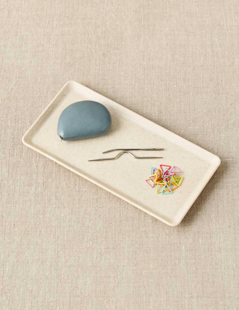 Rectangular white plate with a blue tape measure, two needles, and colorful stitch markers on a beige textured background