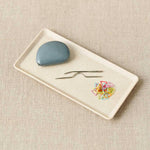 Rectangular white plate with a blue tape measure, two needles, and colorful stitch markers on a beige textured background