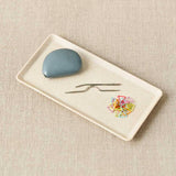 Rectangular white plate with a blue tape measure, two needles, and colorful stitch markers on a beige textured background