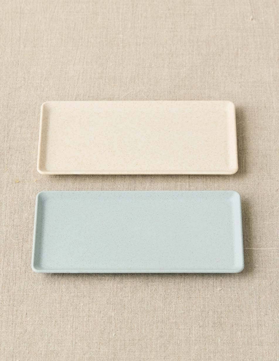 Two rectangular ceramic trays, one beige and one light blue, on a beige fabric background.