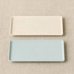 Two rectangular ceramic trays, one beige and one light blue, on a beige fabric background.