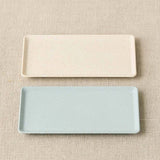 Two rectangular ceramic trays, one beige and one light blue, on a beige fabric background.