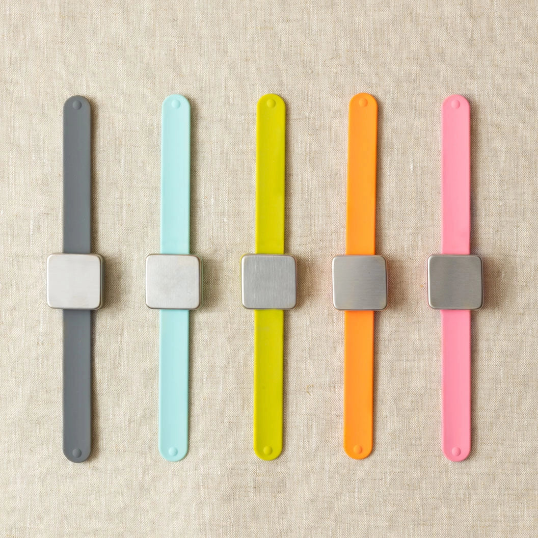 Five colorful wristbands with square magnets for stitch markers on a beige fabric background
