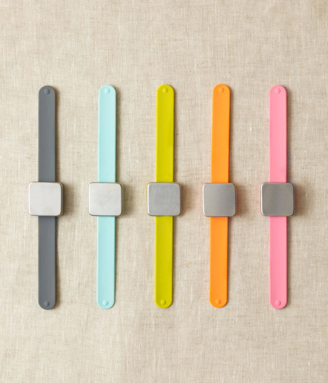 Five colorful wristbands with square magnets for stitch markers on a beige fabric background