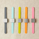 Five colorful wristbands with square magnets for stitch markers on a beige fabric background