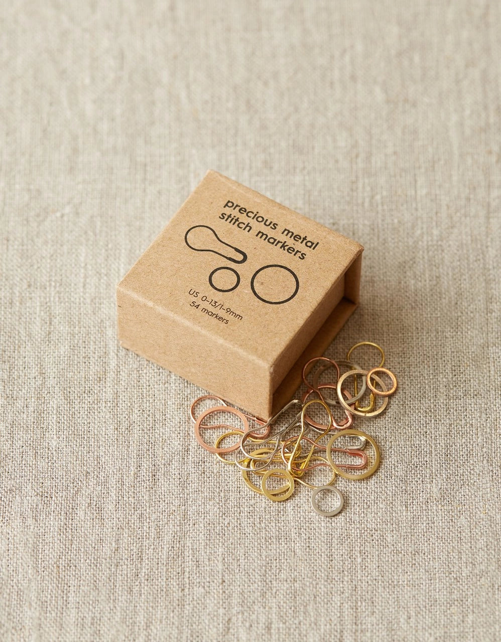 Box of precious metal stitch markers on a textured beige surface
