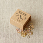 Box of precious metal stitch markers on a textured beige surface