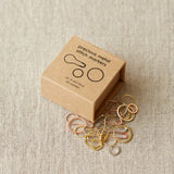 Box of precious metal stitch markers on a textured beige surface