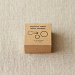 Box of precious metal stitch markers on a beige fabric background